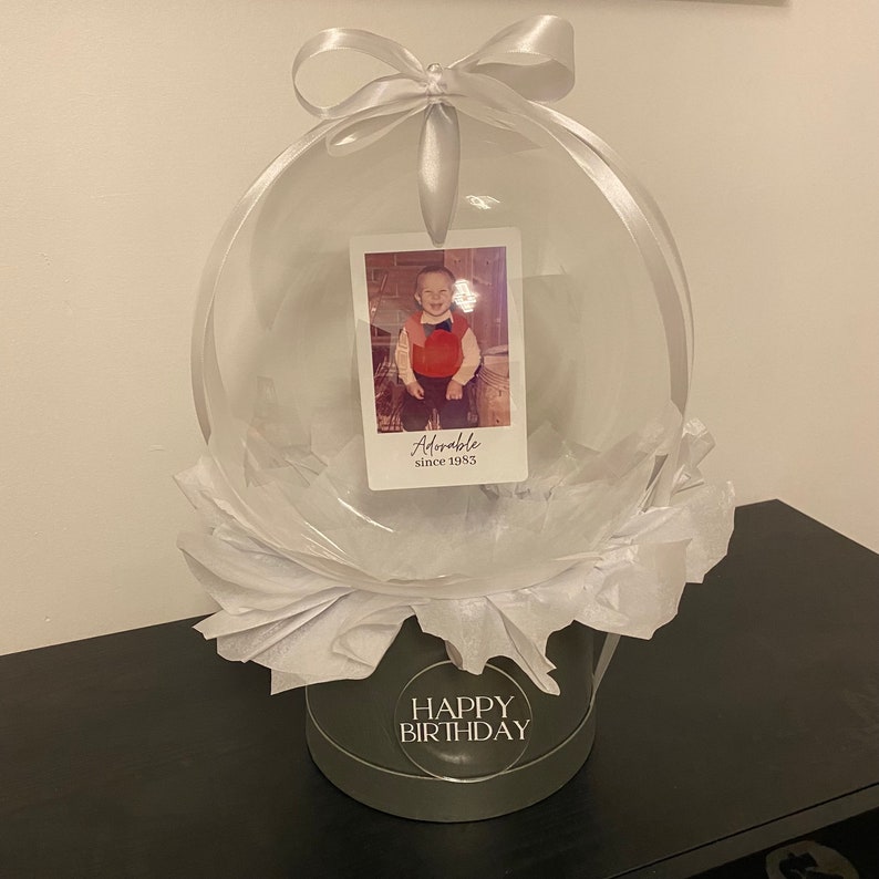 May include: A clear balloon with a white ribbon and a photo inside. The photo shows a young boy wearing a red shirt and a bow tie. The balloon is sitting on a white tissue paper base with a black and white label that says "Happy Birthday".