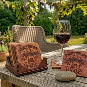 Puede incluir: Posavasos de madera con el texto "Partners in Wine" y un diseño de copa de vino. Una copa de vino tinto está junto a los posavasos. La escena está ambientada al aire libre sobre una mesa de madera.