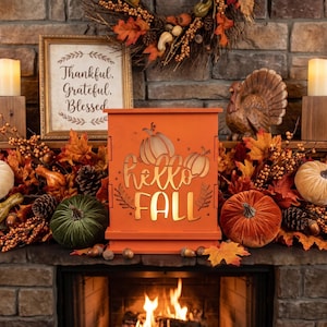May include: An orange rectangular wooden box with a cutout design featuring two pumpkins and the text Hello Fall. The box is surrounded by autumn-themed decor including velvet pumpkins in white, green, and orange, fall leaves, and berries.