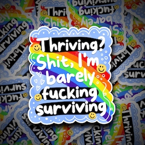 以下が含まれることがあります： 青いスカラップの縁取りと、虹の上に白で「Thriving? Shit, I'm barely fucking surviving」と書かれたステッカー。スマイリーフェイスとハートがテキストの周りに散らばっています。