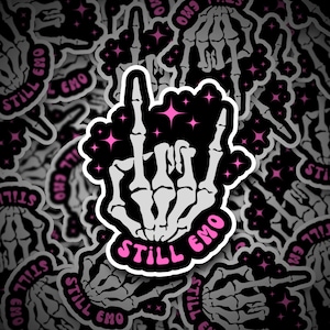 May include: A black and white sticker with a skeleton hand giving the middle finger. The hand is surrounded by pink stars and the text "Still Emo" in a pink circle.