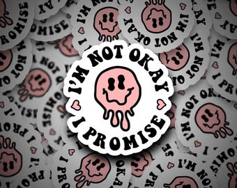 May include: White sticker with the text "I'M NOT OKAY, I PROMISE" in black, surrounding a pink, melting smiley face. Small heart icons are scattered around the edge of the sticker. The stickers are scattered on a white surface.