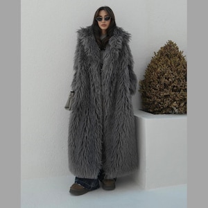 May include: A long, grey faux fur coat with a high collar. The coat is made of soft, fluffy material and has a relaxed fit.