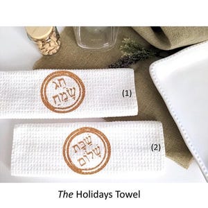May include: Two white waffle-weave kitchen towels featuring circular copper-colored glitter designs. The top towel displays the Hebrew text "Chag Sameach" and the bottom towel displays the Hebrew text "Shabbat Shalom" within the circular borders.