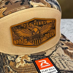 May include: A flat bill snapback hat with a camouflage pattern on the bill and a tan mesh back. The front features a tan patch with the text "250 YEARS AMERICA 2026" and an eagle graphic. The hat also has the Richardson logo.