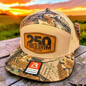 May include: A tan and camouflage flat bill snapback hat with the text "250 Freedom Est. 1776" on a leather patch. The hat has a mesh back and a Richardson tag.