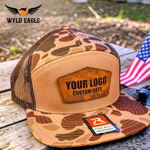 May include: A brown and tan camouflage flat bill snapback hat with a mesh back. The hat features a leather patch with the text "YOUR LOGO CUSTOM HATS". A small American flag and sunglasses are in the background.