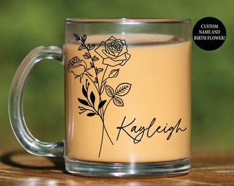 May include: A clear glass mug filled with a light brown liquid. The mug features a black floral design with roses and leaves, along with the name "Kayleigh" in script. A black circle with white text reads "CUSTOM NAME AND BIRTH FLOWER!"