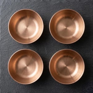 May include: A set of four circular bowls made of polished copper with a metallic finish. Each bowl features a smooth, rounded interior and a consistent, reflective surface texture, arranged in a two-by-two grid pattern for a uniform presentation.