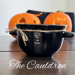 Może przedstawiać: Czarny ceramiczny kociołek z uchwytami, związany sznurkiem, z etykietą z napisem "The Cauldron". Dwie rzeźbione pomarańczowe dynie w tle, sugerujące motyw Halloween.