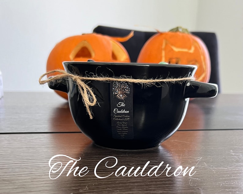 Może przedstawiać: Czarny ceramiczny kociołek z uchwytami, związany sznurkiem, z etykietą z napisem "The Cauldron". Dwie rzeźbione pomarańczowe dynie w tle, sugerujące motyw Halloween.