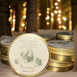 Op de afbeelding: Een stapel goudkleurige, ronde tinnen kaarsen met deksels. De etiketten tonen tekst en een aquarelillustratie van dennenbomen. De tekst luidt "MINI TIN WOODEN WICK CANDLE COCO BEESWAX GROUNDING PINES WALK 3.0 OZ." De merknaam "Spiritual Couture Collection LLC" is ook zichtbaar.