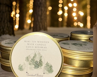 May include: A stack of gold-colored, round tin candles with lids. The labels feature text and a watercolor illustration of pine trees. The text reads "MINI TIN WOODEN WICK CANDLE COCO BEESWAX GROUNDING PINES WALK 3.0 OZ." The brand name "Spiritual Couture Collection LLC" is also visible.
