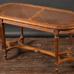 May include: A wooden oval-shaped bench with a woven cane seat and intricate carvings on the legs and frame.