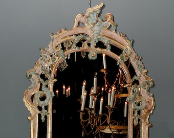 May include: A large, ornate, antique mirror with a carved, gilded frame. The frame is painted in shades of pink, green, and gold. The mirror is reflecting a chandelier with lit candles.