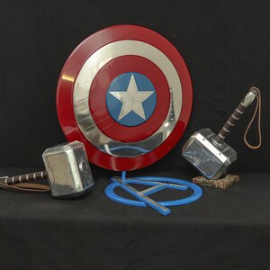 May include: A Captain America shield with a red, white, and blue design, a silver hammer with a brown handle, and a blue Avengers logo. The shield is round with a star in the center. The hammer has the word "Thor" on it.
