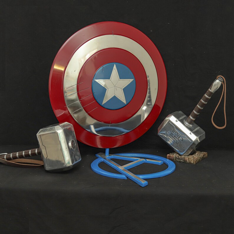 May include: A Captain America shield with a red, white, and blue design, a silver hammer with a brown handle, and a blue Avengers logo. The shield is round with a star in the center. The hammer has the word "Thor" on it.