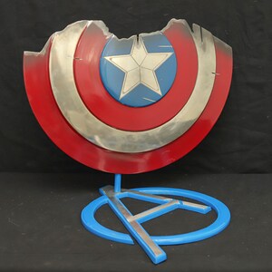 May include: A damaged Captain America shield with a red, white and blue design is displayed on a blue stand with a silver Avengers logo.
