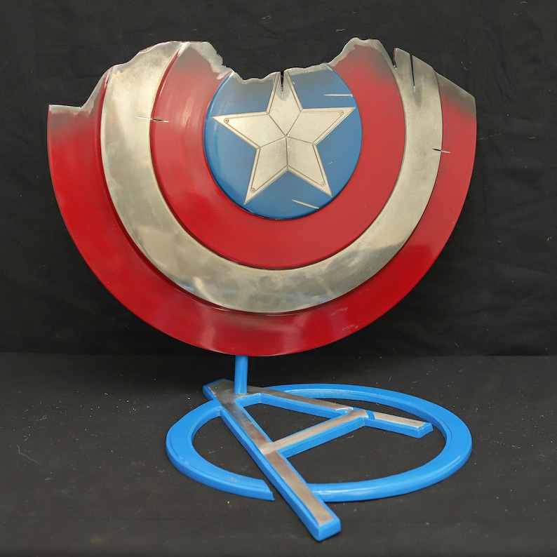 May include: A damaged Captain America shield with a red, white and blue design is displayed on a blue stand with a silver Avengers logo.