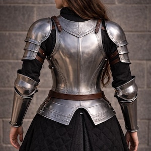 May include: A silver-colored, medieval-style armor set. The armor includes a breastplate, shoulder guards, and arm guards, all connected with brown leather straps. The armor is worn over a black outfit, with a quilted skirt. The armor has decorative etched patterns.