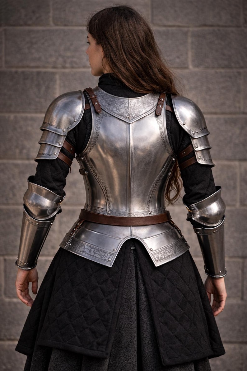 May include: A silver-colored, medieval-style armor set. The armor includes a breastplate, shoulder guards, and arm guards, all connected with brown leather straps. The armor is worn over a black outfit, with a quilted skirt. The armor has decorative etched patterns.