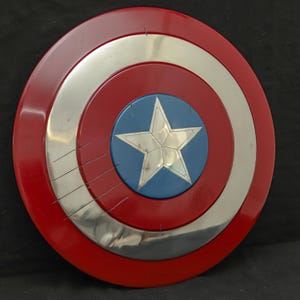 May include: A round shield with a red outer ring, a silver middle ring, and a blue center with a silver star. The shield has a glossy finish and appears to be made of metal. The shield has several scratches on the silver ring.