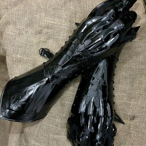 May include: Pair of black, articulated gauntlets with a glossy finish. The gauntlets feature intricate detailing, including stylized claw-like fingers and decorative patterns. A buckle secures the forearm section. The armor is displayed on a textured, neutral-toned surface.