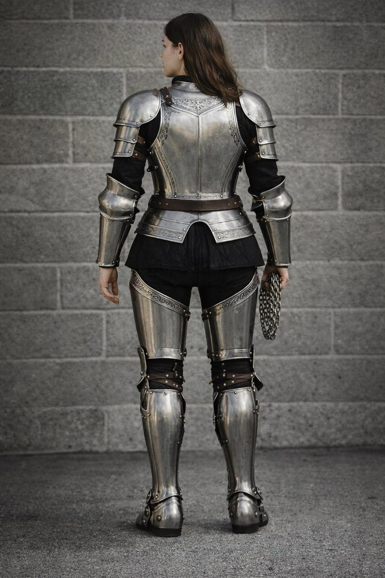 May include: A full suit of silver-colored medieval armor, including a breastplate, pauldrons, arm and leg protection, and boots. The armor is worn over a black outfit. A chainmail is held in the right hand. The background is a gray brick wall.