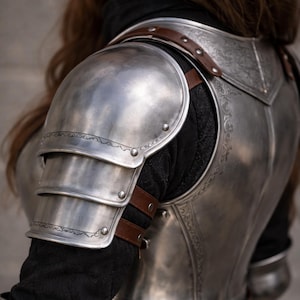 May include: Close-up of a silver metal shoulder armor set with brown leather straps. The armor has decorative etched patterns and is worn over a black garment. The armor is designed to protect the shoulder and upper arm.