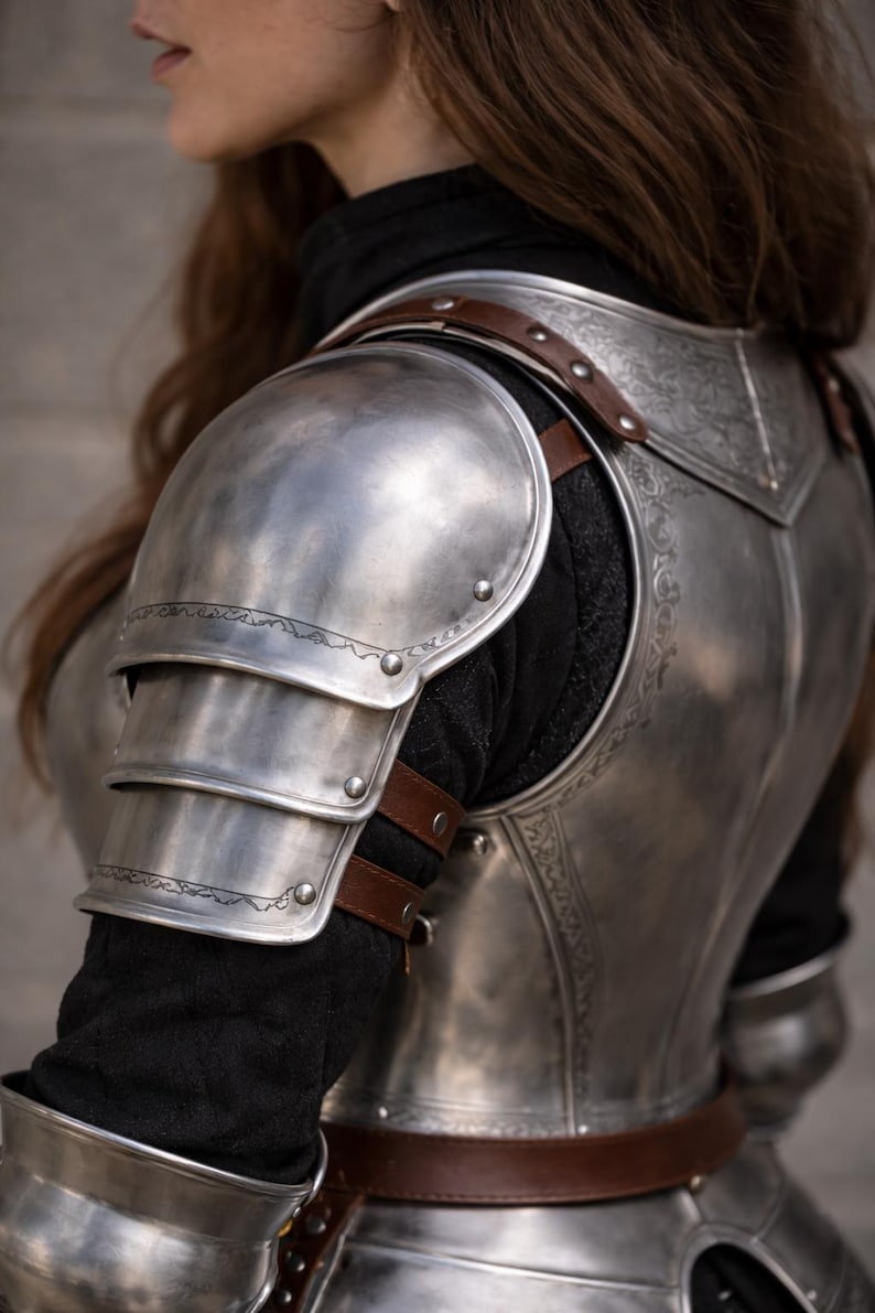 May include: Close-up of a silver metal shoulder armor set with brown leather straps. The armor has decorative etched patterns and is worn over a black garment. The armor is designed to protect the shoulder and upper arm.