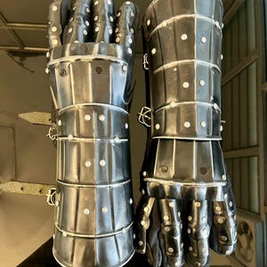 May include: Pair of medieval-style gauntlets made of riveted metal plates. The gauntlets are dark gray with white rivets and feature articulated fingers and wrist protection. The interior appears to be lined with black fabric.