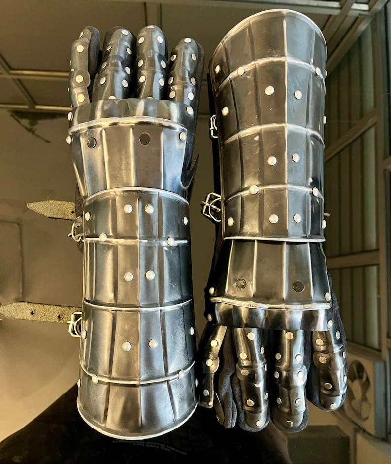 May include: Pair of medieval-style gauntlets made of riveted metal plates. The gauntlets are dark gray with white rivets and feature articulated fingers and wrist protection. The interior appears to be lined with black fabric.