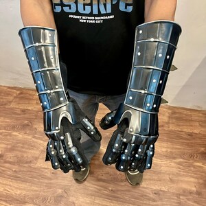 May include: Black metal gauntlets with articulated fingers and wrist protection. The gauntlets are riveted and have a polished finish. The design includes a series of plates and studs, offering a medieval aesthetic. The gauntlets are shown against a neutral background.