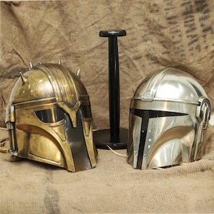 May include: Two Mandalorian helmets, one gold and one silver, are displayed on a black stand. The helmets have a visor and a textured surface.