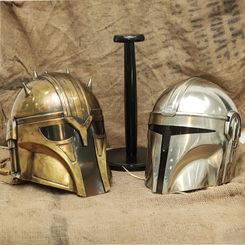May include: Two Mandalorian helmets, one gold and one silver, are displayed on a black stand. The helmets have a visor and a textured surface.