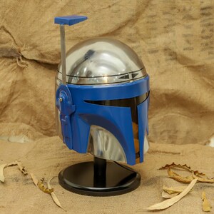 May include: A metallic silver and blue Mandalorian helmet, displayed on a black stand. The helmet features a blue visor and accents, with a silver dome and a blue antenna. The background is a neutral tan.