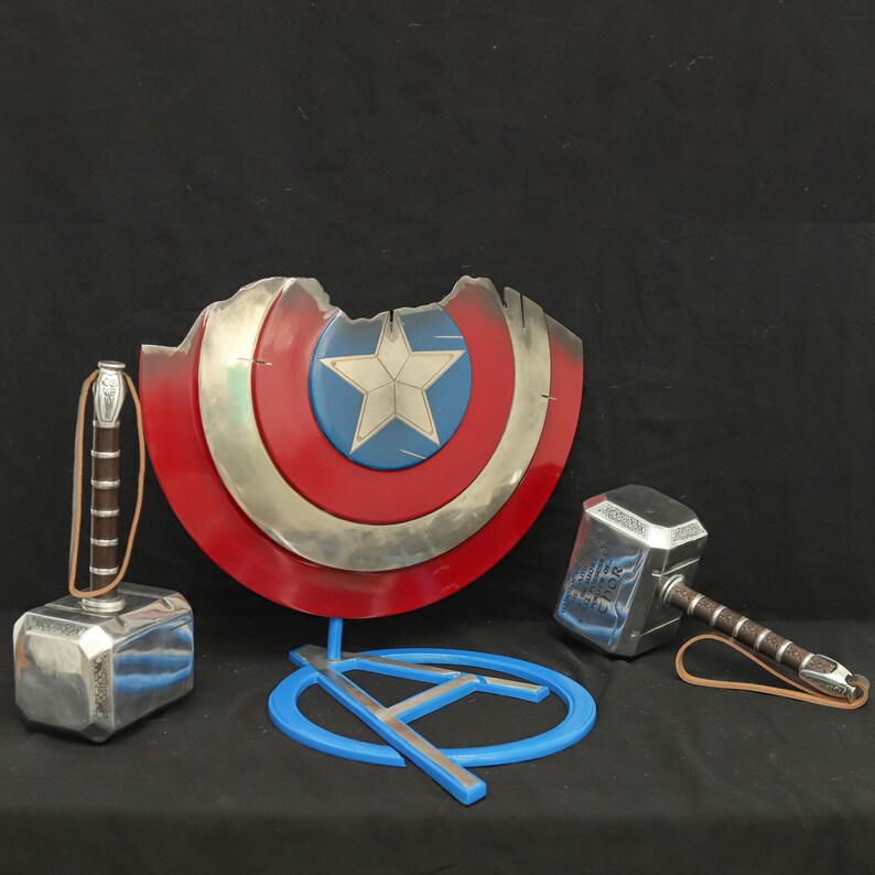 May include: A silver Captain America shield with a red, white and blue star design, a silver Thor's hammer, and a blue stand with a silver A.