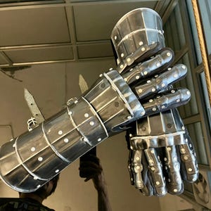 May include: A pair of steel gauntlets, likely for historical reenactment or display. The gauntlets are riveted with white dots and feature articulated fingers and wrist protection. The metal has a dark, reflective finish.