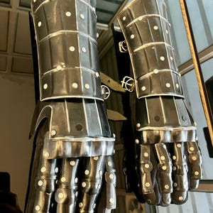 May include: A pair of medieval-style gauntlets, crafted from dark metal plates and rivets. The gauntlets feature articulated fingers and wrist protection, with a dark fabric interior. The design suggests historical armor.