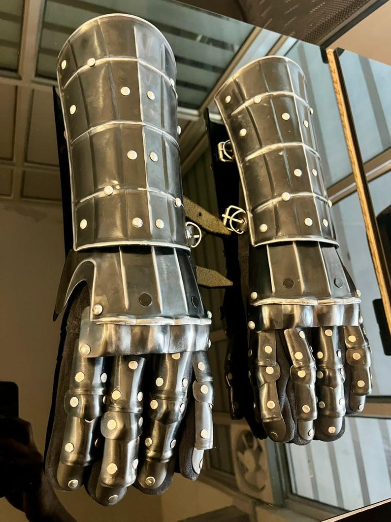May include: A pair of medieval-style gauntlets, crafted from dark metal plates and rivets. The gauntlets feature articulated fingers and wrist protection, with a dark fabric interior. The design suggests historical armor.