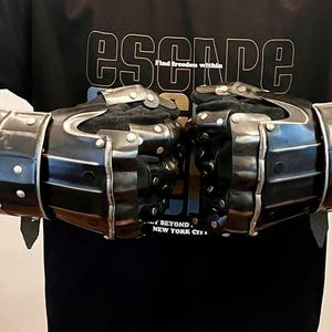 May include: Black metal gauntlets with silver rivets and leather straps. The gauntlets are segmented and designed to protect the hands and forearms. The image also shows a black t-shirt with the word "ESCAPE" printed on it.