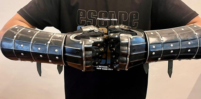 May include: Black metal gauntlets with silver rivets and leather straps. The gauntlets are segmented and designed to protect the hands and forearms. The image also shows a black t-shirt with the word "ESCAPE" printed on it.