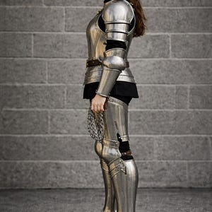 May include: Full suit of polished steel armor, including a breastplate, shoulder guards, arm and leg protection, and boots. The armor is worn over a black garment. A chainmail piece is held in the hand. The background is a gray brick wall.
