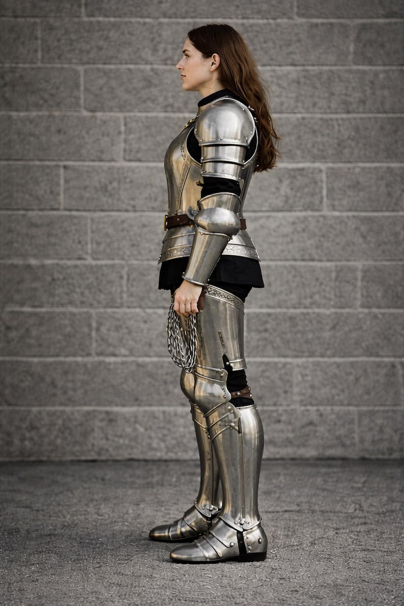 May include: Full suit of polished steel armor, including a breastplate, shoulder guards, arm and leg protection, and boots. The armor is worn over a black garment. A chainmail piece is held in the hand. The background is a gray brick wall.