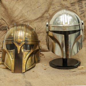 May include: Two helmets, one gold and one silver, are displayed on a brown surface. The gold helmet has a textured surface and a visor. The silver helmet has a smooth surface and a visor.