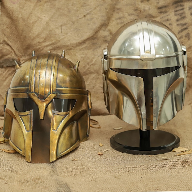 May include: Two helmets, one gold and one silver, are displayed on a brown surface. The gold helmet has a textured surface and a visor. The silver helmet has a smooth surface and a visor.
