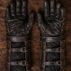 May include: Pair of black leather gauntlets with metal accents and buckle closures. The gloves have a detailed design with rivets and straps, suggesting a medieval or fantasy theme. The gloves are displayed on a wooden surface.