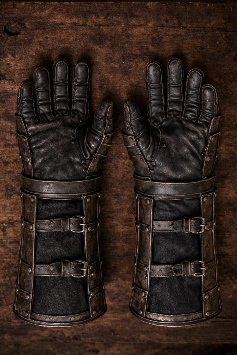 May include: Pair of black leather gauntlets with metal accents and buckle closures. The gloves have a detailed design with rivets and straps, suggesting a medieval or fantasy theme. The gloves are displayed on a wooden surface.