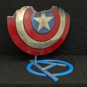 May include: A red, white and blue Captain America shield with a silver star in the centre. The shield is damaged and has a large chunk missing. The shield is on a blue stand with a silver A.