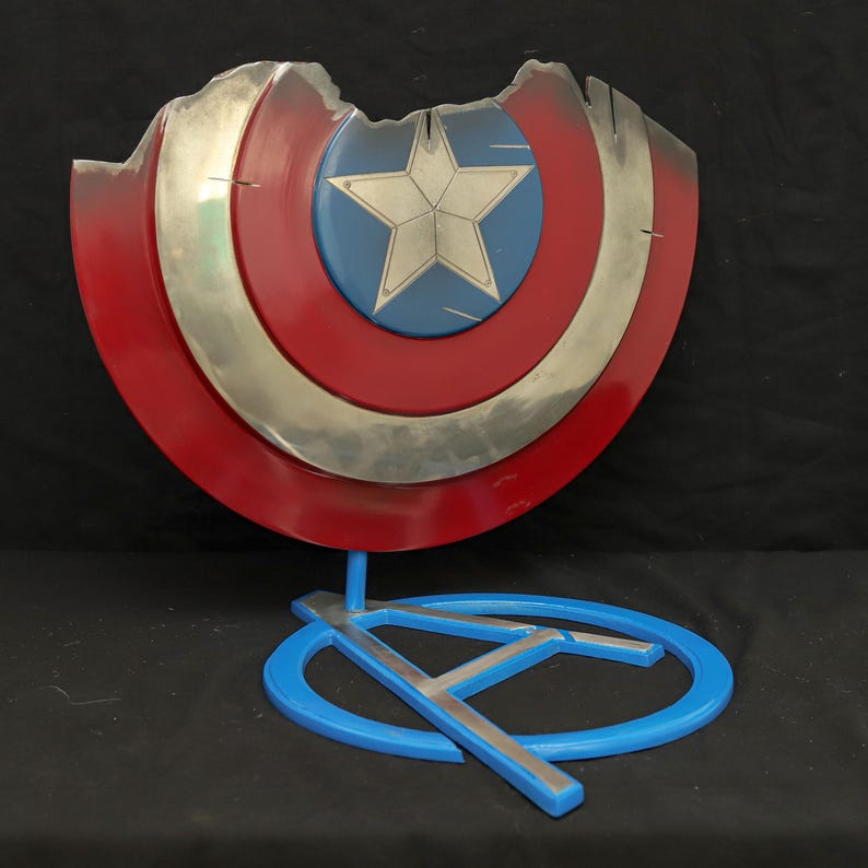 May include: A red, white and blue Captain America shield with a silver star in the centre. The shield is damaged and has a large chunk missing. The shield is on a blue stand with a silver A.